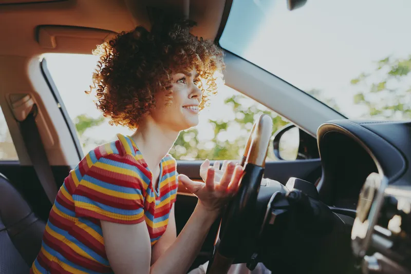 Cheerful woman driving a car, striped shirt, sunlit moment, relaxed posture, candid lifestyle, warm tones, natural light, vibrant greenery, bright outdoor setting, with joyful energy
