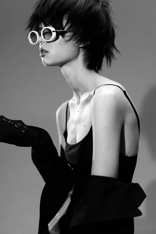 Black and white portrait of a stylish woman wearing sunglasses and a dark outfit with a modern hairstyle and serious expression.