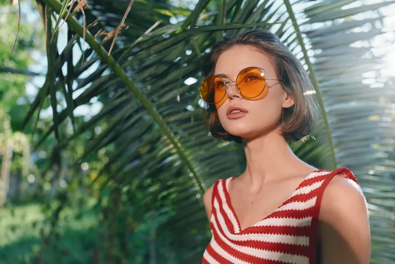 A stylish woman wearing orange sunglasses and a red and white striped top stands outdoors among lush tropical plants, exuding confidence and a carefree sunny summer vibe. Natural light highlights her