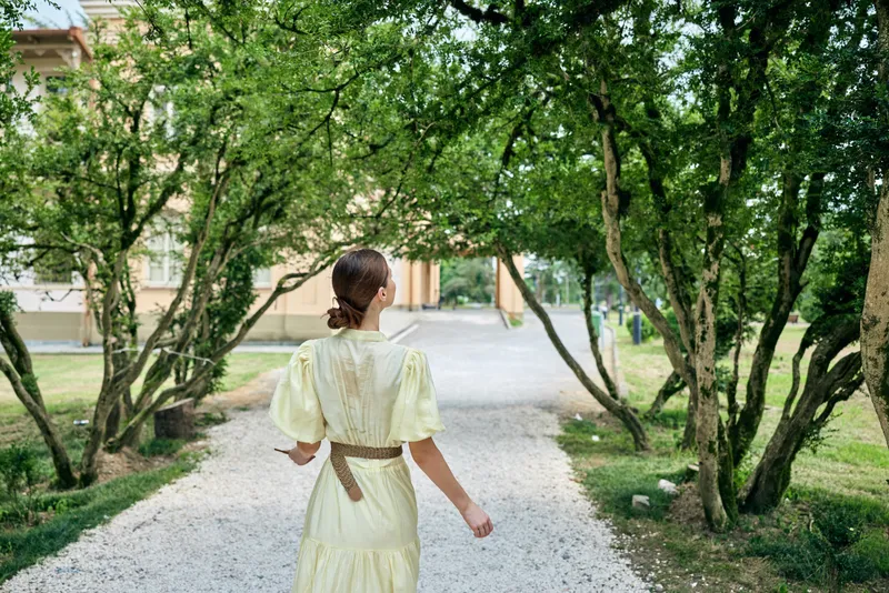 Woman in yellow dress walking on garden path among trees back view summer braid authenticity inclusivity calm serene grace