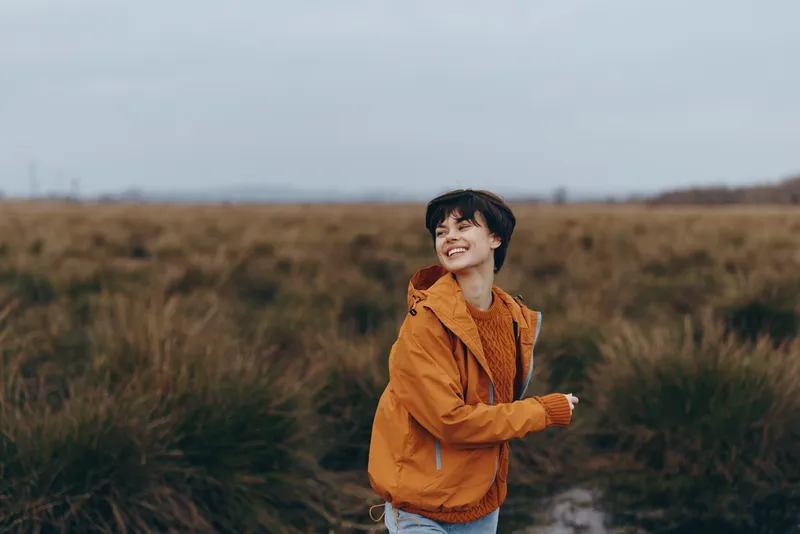 Woman lifestyle outdoors, smiling in a casual orange jacket, standing on a rural trail, surrounded by grass and open field under cloudy sky, calm mood