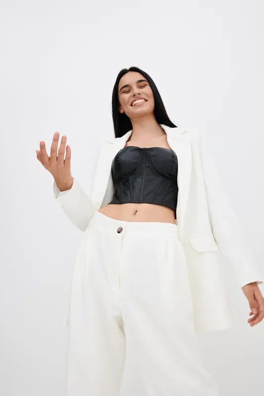 smiling woman in white pants and blazer with black corset top, posing confidently against plain light background, casual modern fashion style
