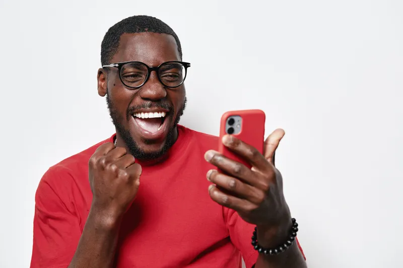 African American man with dark skin wearing glasses and red shirt celebrating with fist pump while looking at smartphone screen isolated on white background with joyful expression.