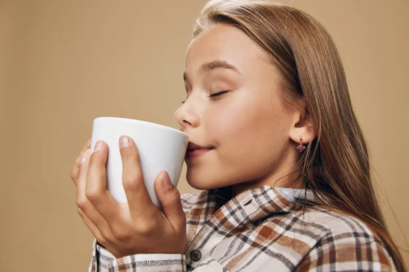 Girl in plaid shirt holding white mug close to face with eyes closed enjoying aroma of hot drink against plain background, cozy and relaxed moment.