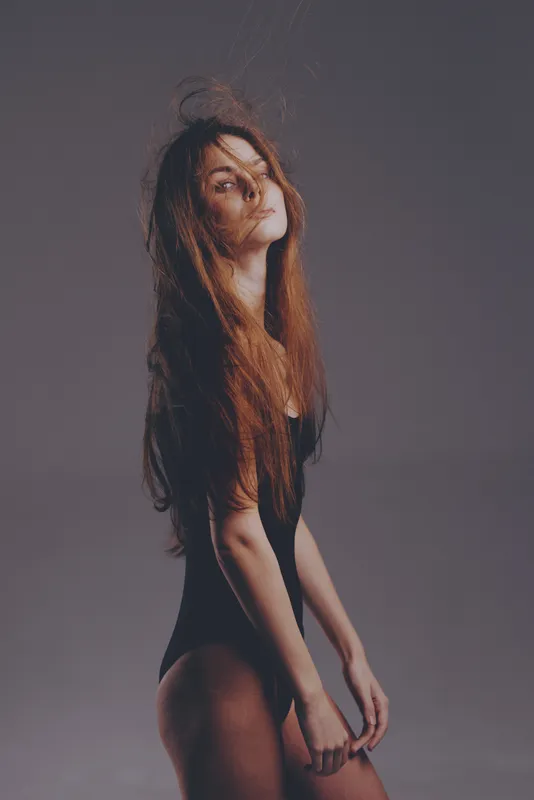 Portrait of a slender woman with long hair in a black bodysuit posing against a gray background with soft lighting.