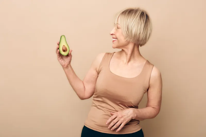 Smiling mature woman with short blonde hair holds half an avocado, looking at it with interest. Concept of healthy lifestyle, nutrition, natural food, and wellness for older adults.