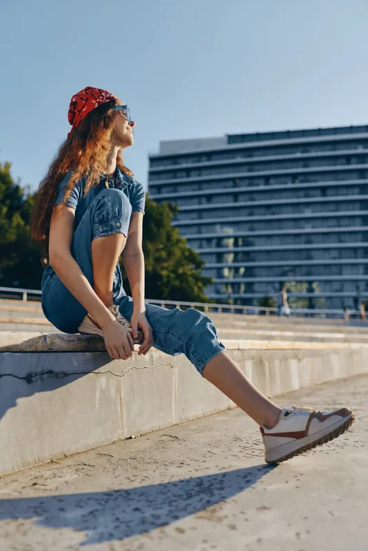 lifestyle woman in modern boho-western denim street style sitting outdoors wearing bandana and casual shoes with sunny urban background in warm film color tone