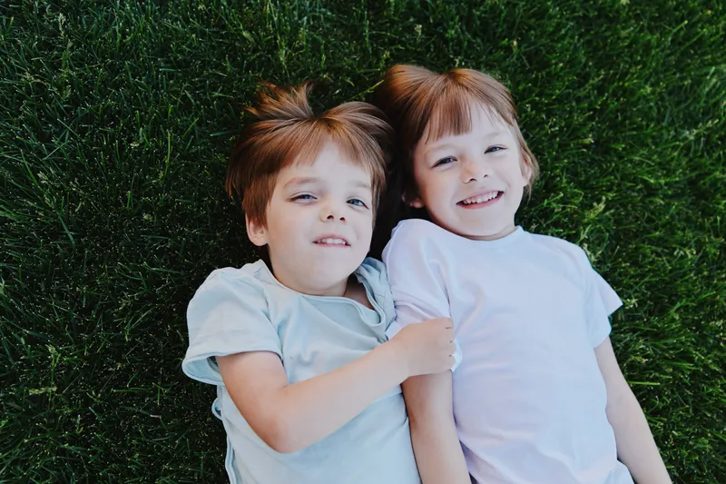 Two cheerful children lying on green grass, enjoying a sunny day outdoors, with bright smiles and playful expressions, creating a joyful atmosphere.