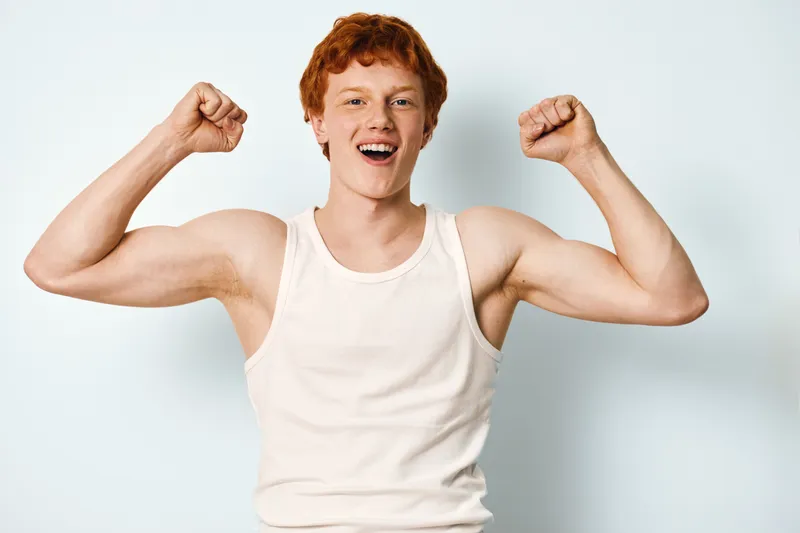 Young man with red hair wearing white tank top flexing muscles and celebrating success with happy facial expression on white background in studio. Positive emotion and strength.