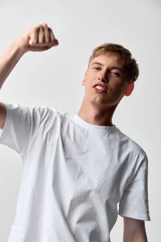 Young man, energetic fist, strong gesture and confident stance in a white tee, studio setup, dynamic pose conveying motivation, victory and bold attitude in a bright minimal environment