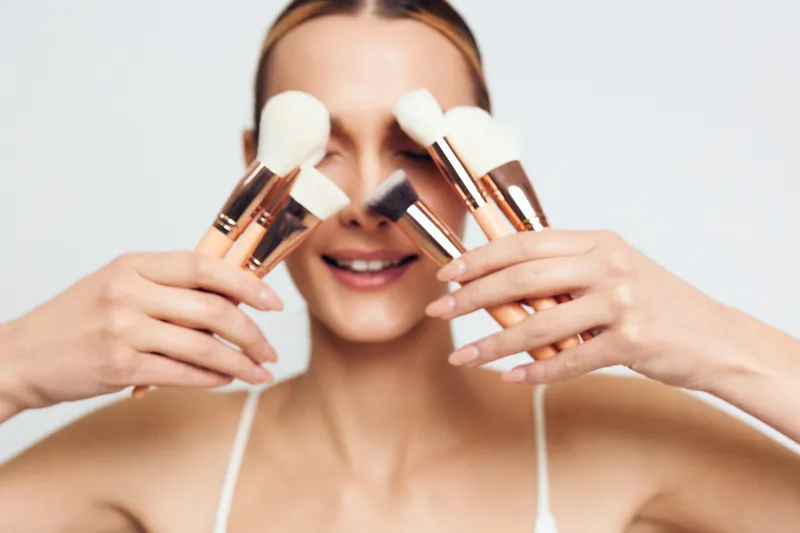 Young woman with blonde hair smiles while holding several white makeup brushes near her face, enjoying a beauty routine in a bright studio setting.