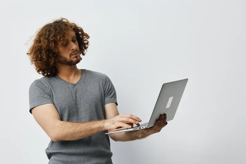 Young man with curly hair working on a laptop, wearing a casual gray t shirt against a plain background, exuding concentration and focus in a modern workspace