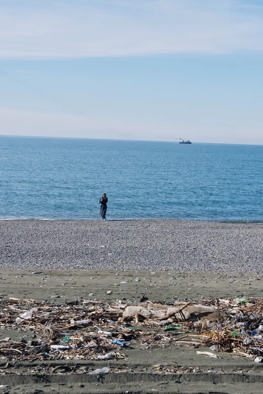 Beach scene with a lone person standing on a pebble shore, distant boat on the horizon, foreground litter and debris highlighting pollution and environmental concerns to highlight pollution.