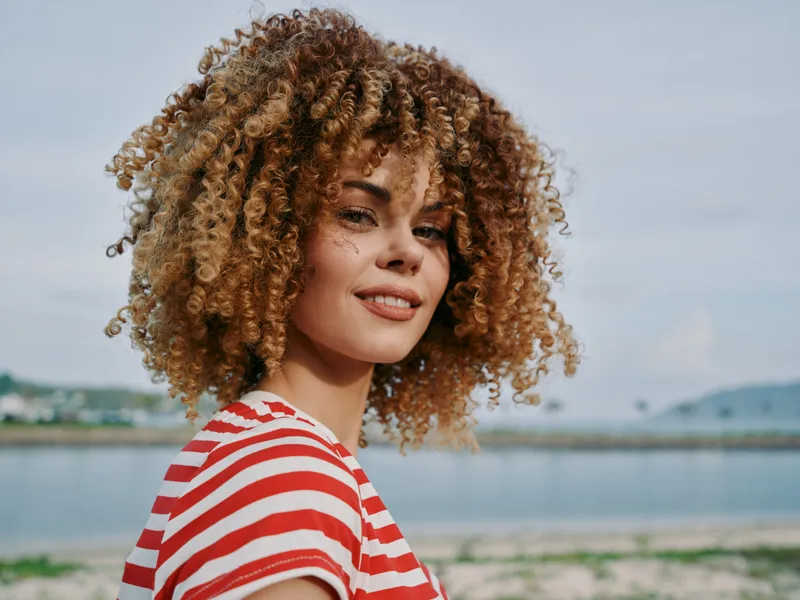 A portrait of a smiling woman with curly hair on a bright beach, wearing a red and white striped shirt, capturing a warm, sunny moment against blue water and wide sky.
