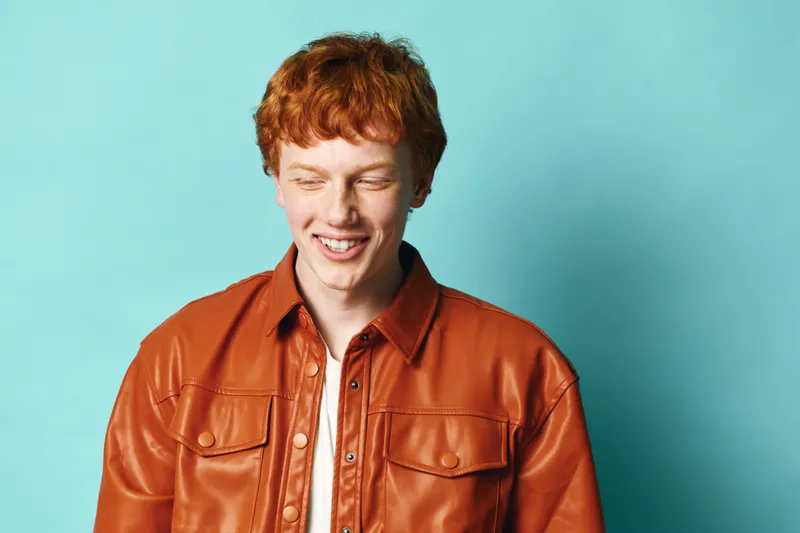 Young man with red hair wearing brown leather jacket smiling with eyes closed against blue background. Portrait of cheerful male adult with casual style isolated indoors.