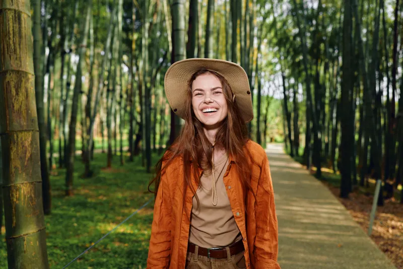 Woman smile hat bamboo forest path, portrait of a happy young female walking outdoors on a sunlit boardwalk, casual jacket and straw hat, cheerful candid lifestyle shot in nature.