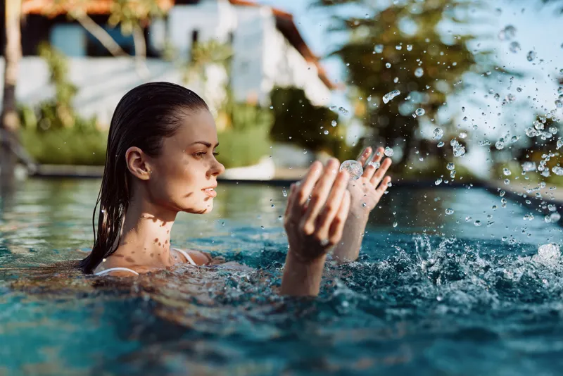 A young woman in a swimming pool splashes water with her hands while enjoying a sunny day outdoors. The background shows blurred trees and buildings under natural light.
