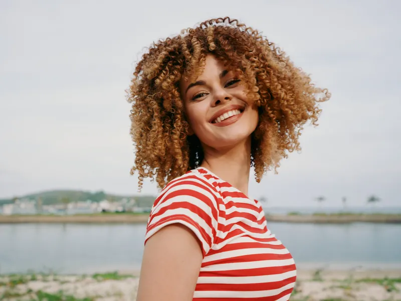 Portrait of a smiling woman with curly hair wearing a red striped shirt, standing outdoors beside a calm waterfront, bathed in bright natural light and confident energy.