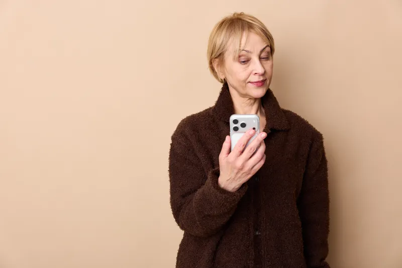 Elderly woman holding smartphone indoors portrait, reading a message with gentle smile, beige background, brown coat, focused eyes, communication technology, calm mood