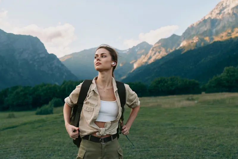 Young woman with a backpack stands confidently in a mountain meadow looking into the distance during a sunny day with clear skies and rugged peaks behind her.