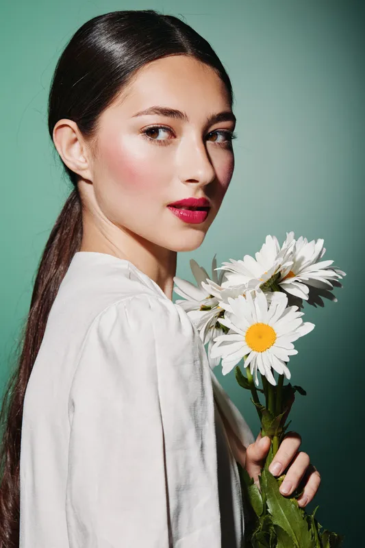 Young woman with sleek ponytail wearing white holds daisies thoughtfully against a pastel green background. Soft lighting enhances her rosy makeup and stylish appearance. Beauty concept.