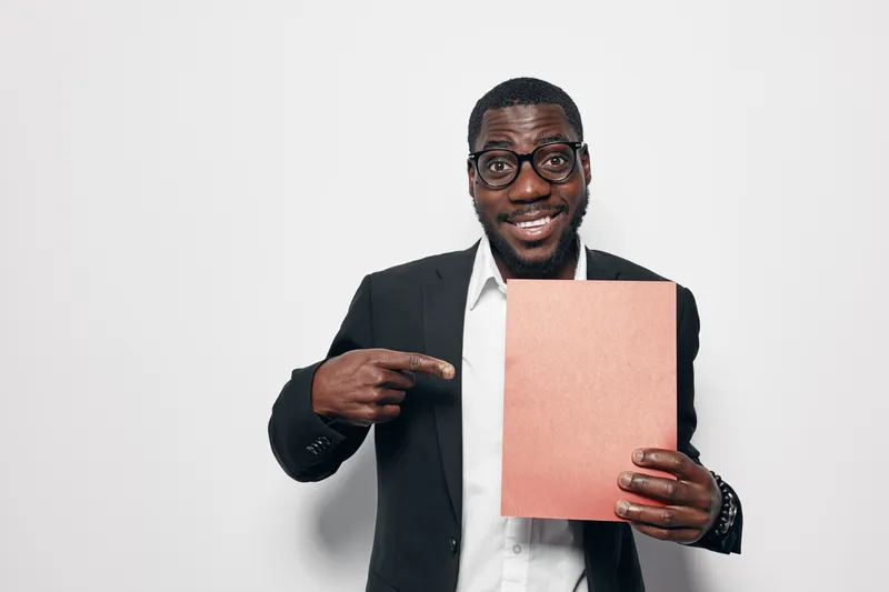 African American man with dark skin and glasses in black suit holding and pointing at blank folder for presentation or business concept on white background.