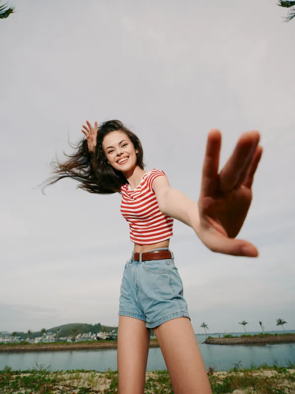 Energetic young woman leans toward the camera with a bright smile, wind in her hair, wearing a striped red top and denim shorts, capturing a vivid outdoor moment.