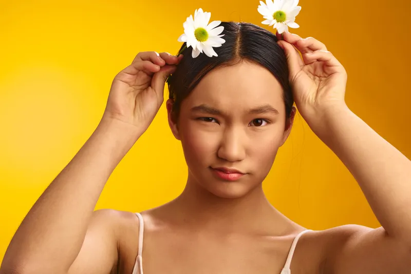 Asian woman stands against a bright yellow backdrop, wearing a light top and floral headpieces. She touches daisies with a calm, confident gaze, conveying freshness, creativity, and a sunny mood.