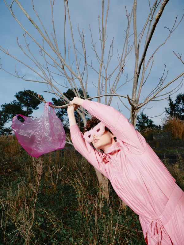 Person in pink dress outdoors, reaching for a bag, playful pose among bare trees on a sunny day, expressive fashion moment and joyful movement in nature.