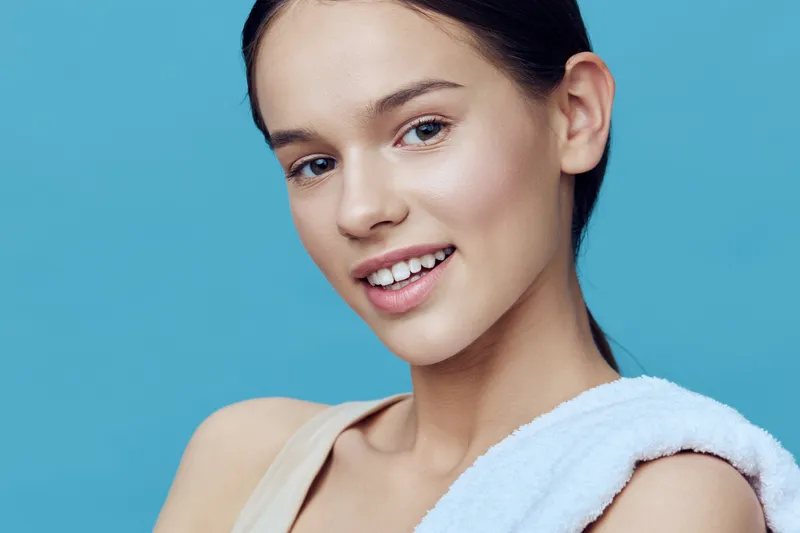 Young woman with a fresh, radiant smile showcasing healthy skin, wearing minimal makeup on a soft blue background, radiating positivity and beauty