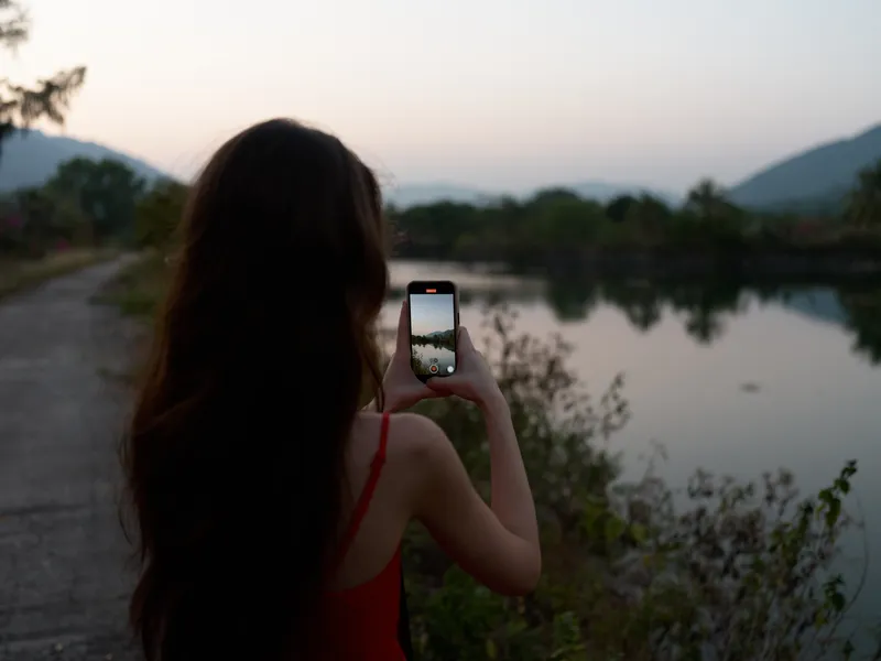 young woman with long hair taking photo of lake and mountains with smartphone during sunset, back view, peaceful nature scene with soft evening light