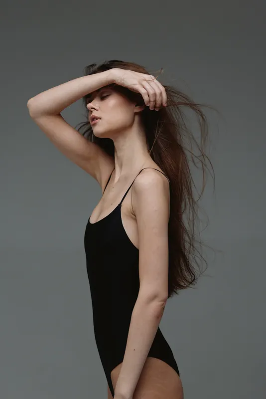 Elegant woman in black bodysuit posing with eyes closed against neutral gray background, showcasing calm and graceful style.