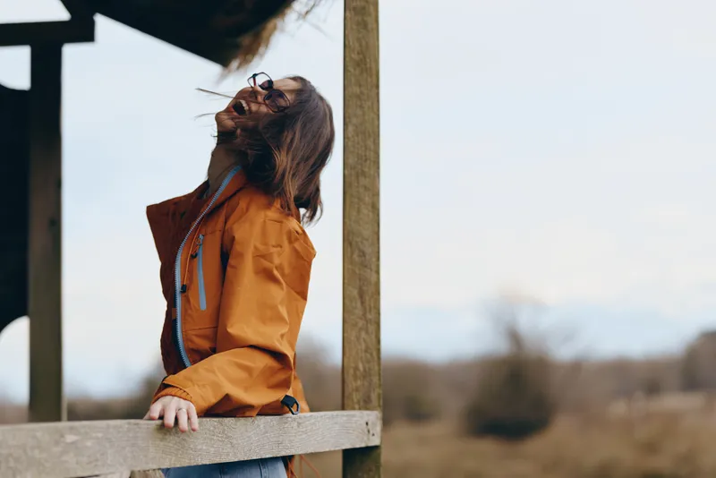 Woman in orange jacket outdoors, paused during walking in park, leash in hand as a dog stands close on a wooden railing, lifestyle portrait, soft daylight, calm