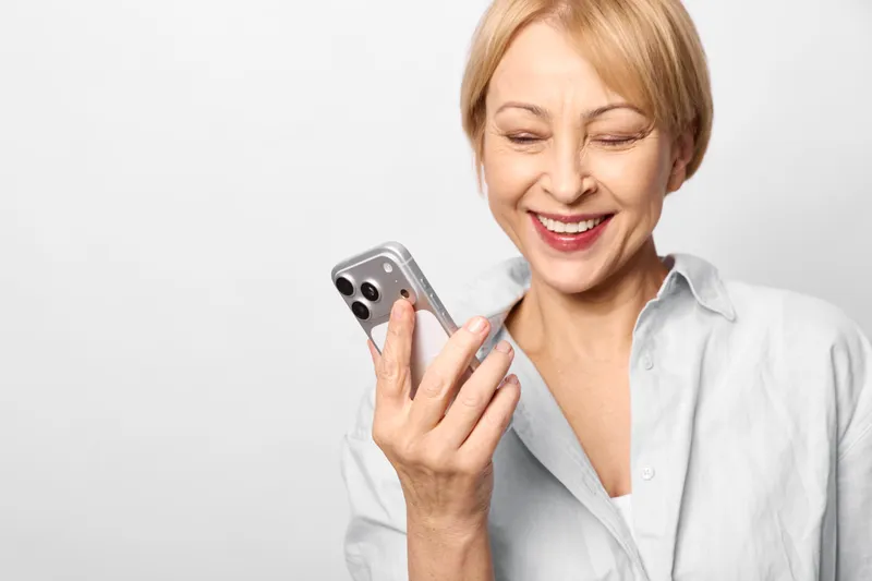 Woman smartphone happy portrait laughing elderly female, holds mobile phone and smiles while chatting, indoor white background, closeup, casual shirt, joyful expression