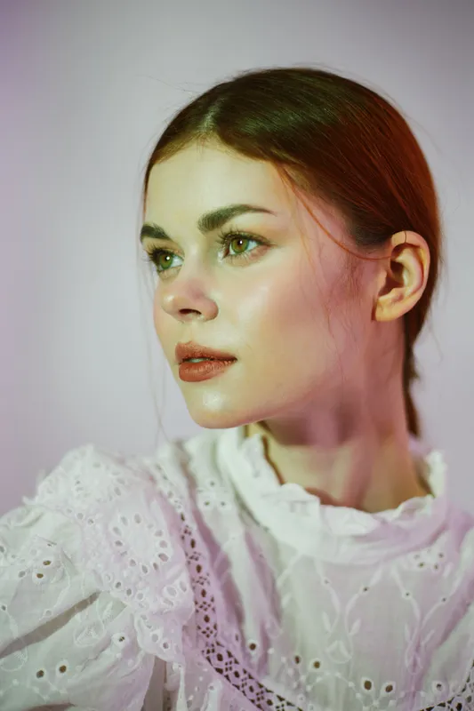 Elegant young woman with green eyes and light skin, wearing a white lace blouse, showcasing a serene expression against a soft, pastel background