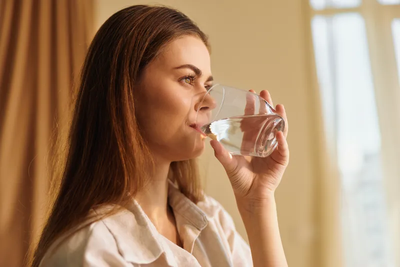Woman drinking water from a glass in a morning home portrait, relaxed and thoughtful with warm natural sunlight, healthy hydration routine and calm indoor lifestyle moment.
