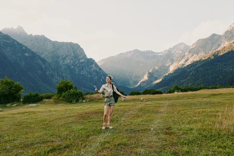 Young woman with long hair walks across a green meadow surrounded by mountains during a sunny day, enjoying nature and outdoor adventure with a casual outfit.