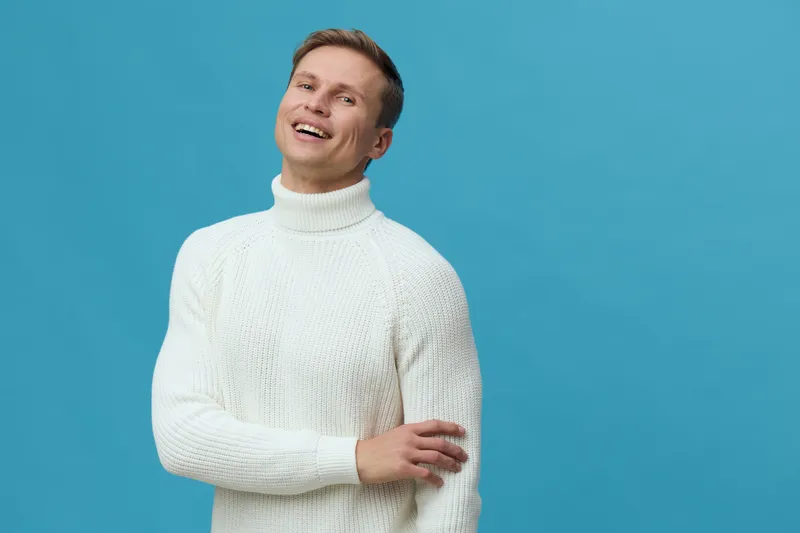 Smiling man in white turtleneck sweater looks joyful and relaxed against a bright blue background, conveying happiness, positivity, friendliness, warmth, casual style, and confidence. Studio portrait 