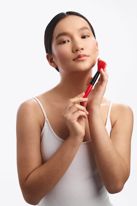 Asian woman applies makeup with a soft brush on her cheek in a clean studio setting. She gazes slightly away, exuding calm confidence and a fresh, natural look enhanced by gentle lighting and a