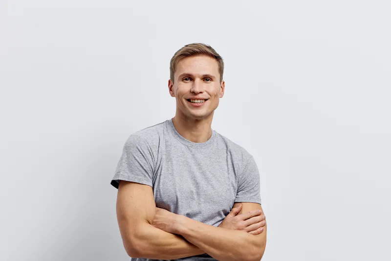 Smiling man with crossed arms wearing casual grey t-shirt, looking confidently at camera, isolated on plain white background. Studio portrait, relaxed mood, male model, happy expression, everyday clot