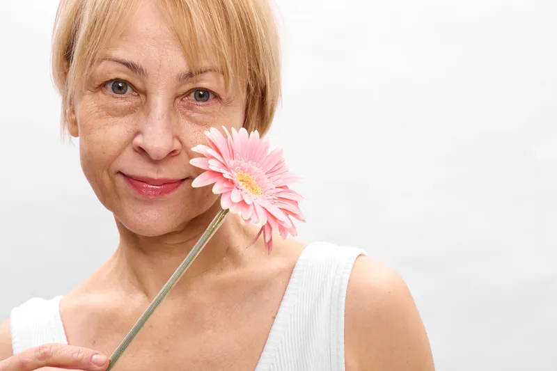 Blonde woman portrait holding a pink flower close to her face, soft smile, calm expression, white background, daylight, beauty, skincare, healthy skin, spring mood