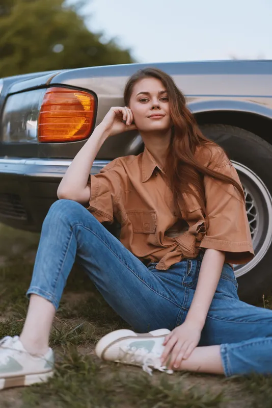 Young woman sitting casually next to a vintage car, wearing a brown oversized shirt and jeans, exuding a relaxed vibe perfect for fashion and lifestyle photography.