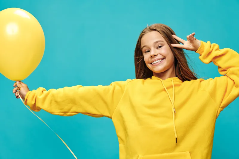 Young girl smiling and holding yellow balloon with hand making peace sign against blue background wearing casual hoodie and looking cheerful and happy.