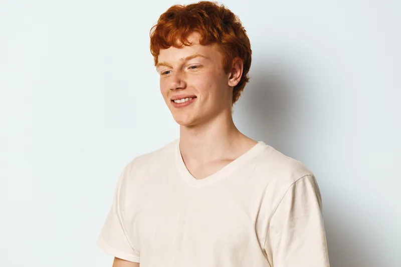Young man with red hair wearing white t shirt stands smiling looking down on plain white background. Casual style, happiness, portrait and relaxed mood in studio setting.