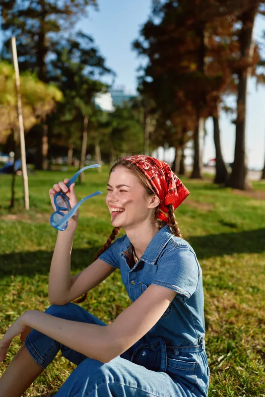 lifestyle woman in modern boho-western denim street style laughing outdoors, with film color effect, wearing red bandana and holding blue sunglasses in sunny park setting