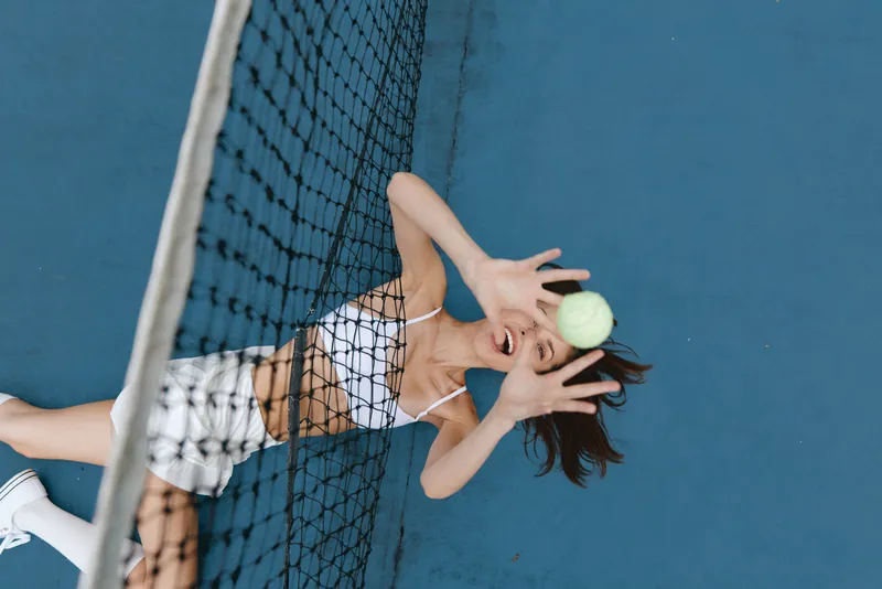 Joyful young woman catching a tennis ball during a game on a blue court, wearing sporty attire and radiating happiness