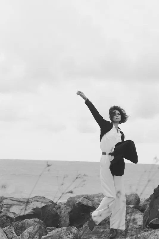 Woman on a rocky coast strikes a confident pose as wind lifts her jacket, monochrome scenery capturing fashion, motion, outdoor mood, and seaside elegance against rough stones, dramatic contrast and