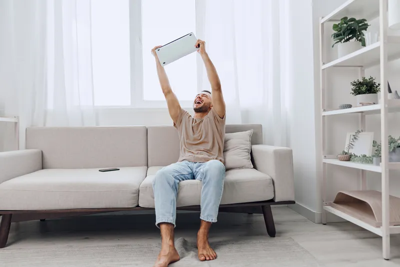Happy man celebrating success at home, holding a laptop above his head. The atmosphere reflects joy and triumph, capturing the essence of modern remote work.