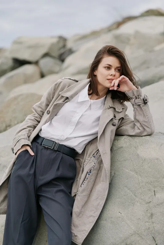 Young woman in textured coat and white shirt leans on rock outdoors, looking sideways with thoughtful expression. Casual fashion, nature, and relaxed mood in natural light.