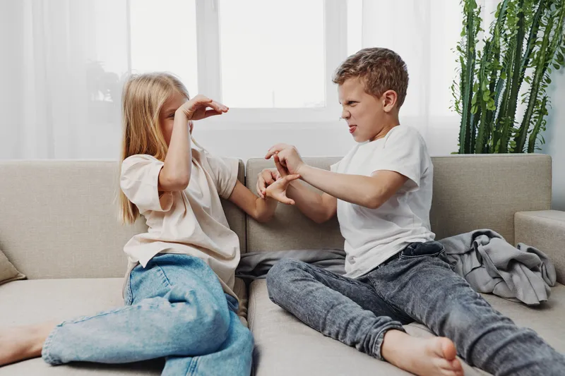 Two children sitting on sofa playfully fighting with expressive faces in bright living room. Concept of sibling rivalry, childhood emotions, and family dynamics.